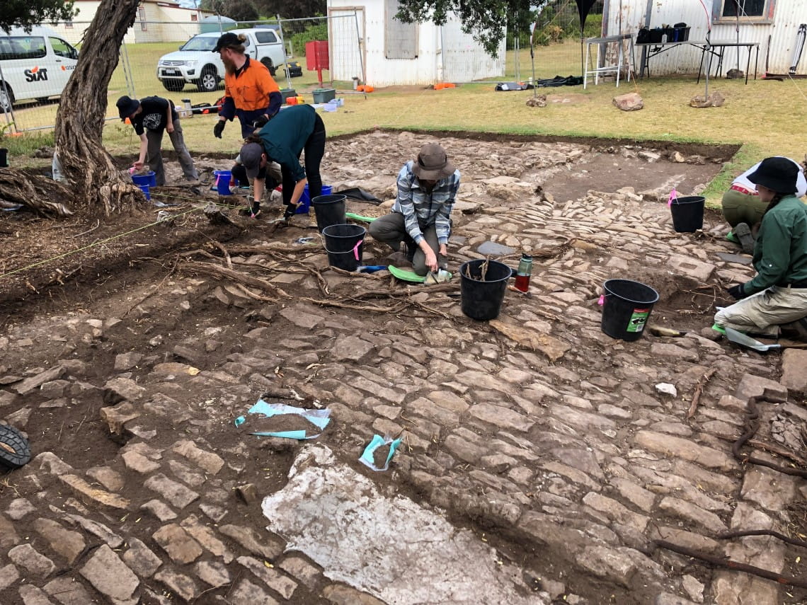 A group of people uncovering a cobbled pavement from beneath a green lawn.