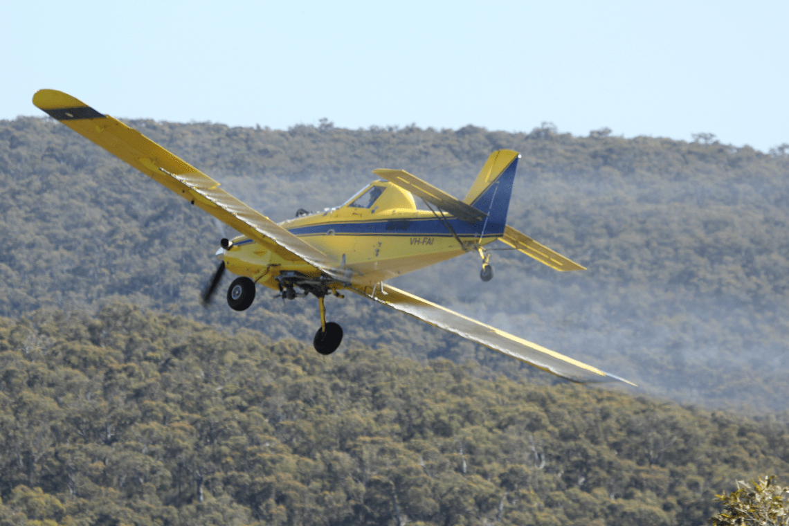 A yellow plane flying over Great Otway National Park. It is tilted to the right and the phosphite looks like a fine mist falling towards the right hand side of the park.