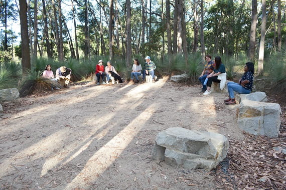 Large stones arranged in a semicircle around a large open area and surrounded by trees. Diverse people are sitting around on the stones conversing and laughing together. 