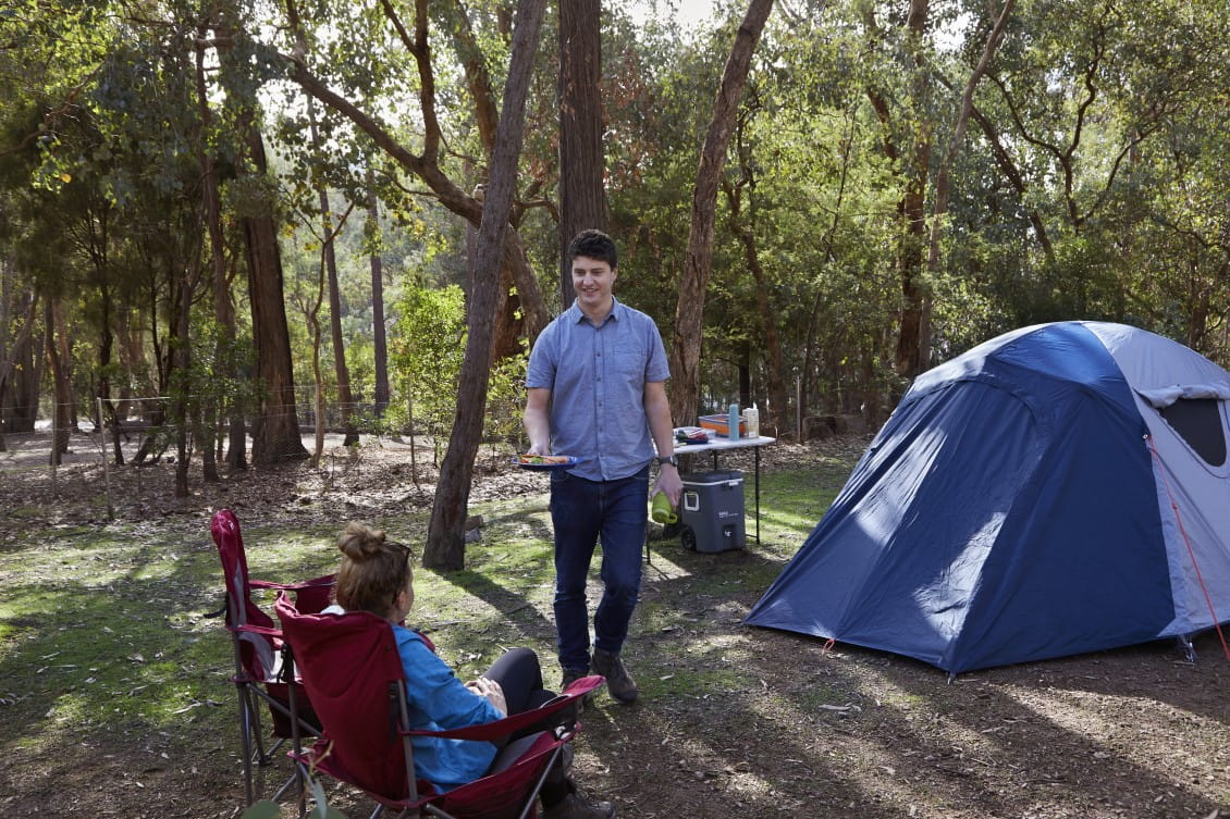 A man stands next to a tent, holding a plate of food. A woman sites in a chair next to him.