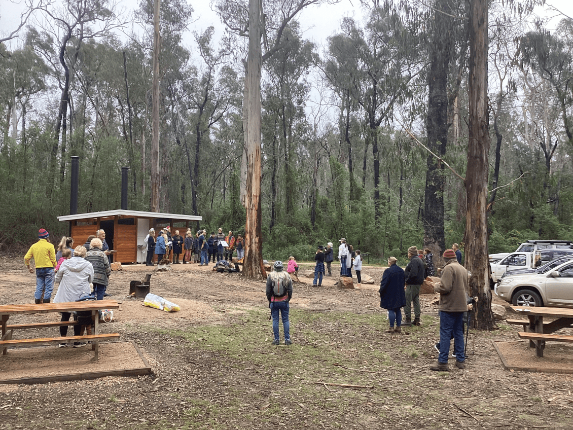 A group of people gathered in a forest clearing with picnic tables. Some of the trees show burn marks from bushfires.