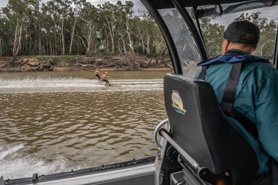 Image shows a Parks Victoria staff member in a boat watching two people water skiiing on the river. Fallen trees and branches on the riverbank are in the background.