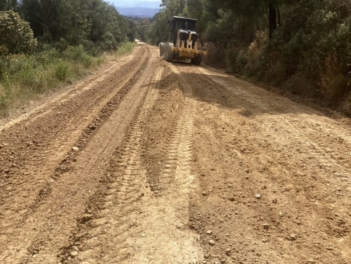 A road-grading machine drives down a dirt road spreading gravel