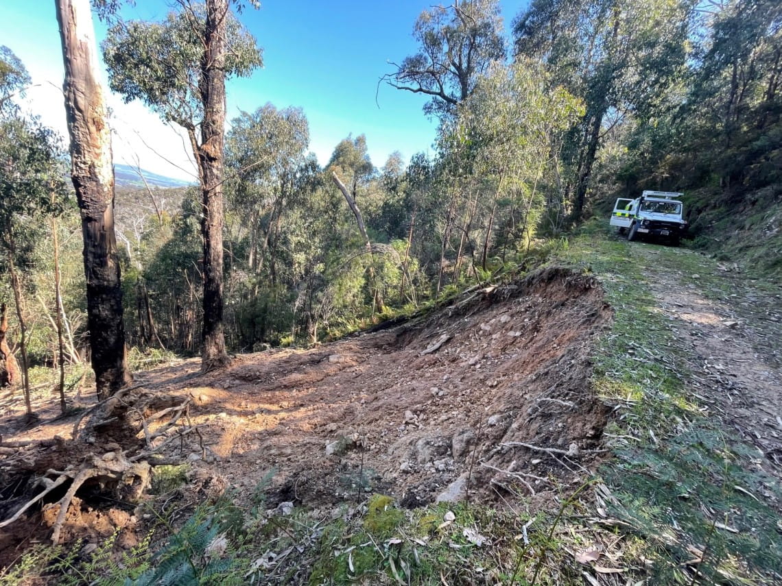 A car is at the top of a road, overlooking a landslip which has severely damaged the surrounding road and incline in Kinglake National Park