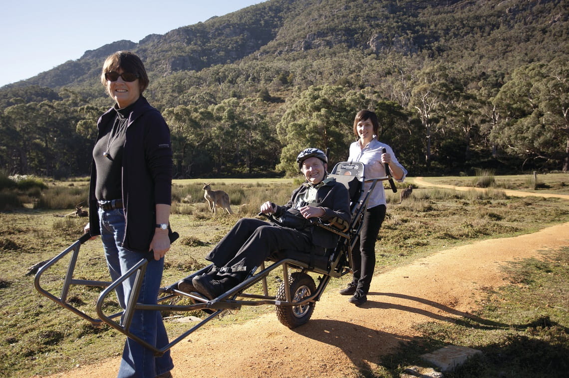 Two people assisting male in an accessible Trail Rider with kangaroo in background
