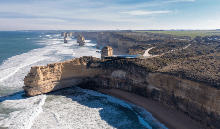 A wide aerial view of a rugged coastline along the Great Ocean Road, showing towering limestone cliffs and several offshore rock stacks rising from the ocean. White waves roll toward a narrow sandy beach below, while a modern viewing platform sits on the cliff edge overlooking the landscape under a clear blue sky.