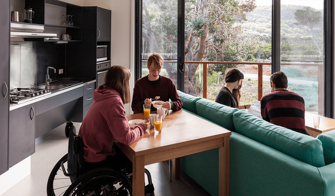 A woman in a wheelchair and a male eat breakfast at a table while a male and female sit on a couch