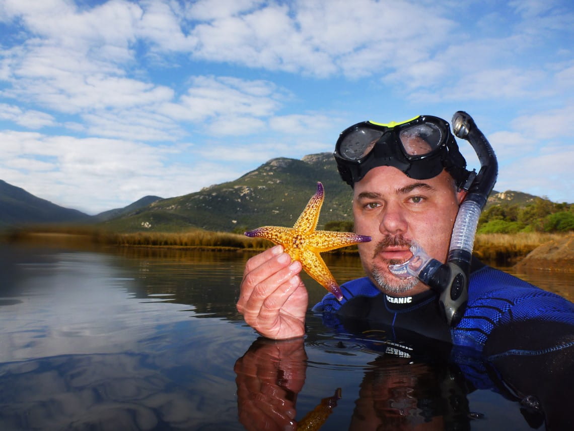 A man in diving gear, up to his neck in water, holds up a starfish