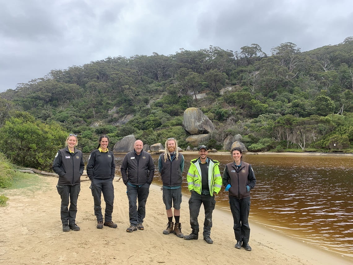 Six smiling people in Parks Victoria uniforms standing in a line on a river bank, with brown water and a scrub-and-boulder-covered hill behind them (Tidal River, Wilsons Promontory National Park)