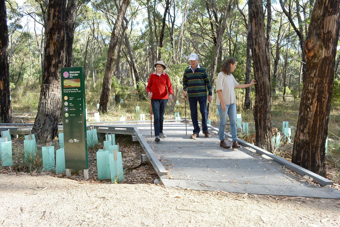 An elderly couple walk along a raised walking track. A woman near them is touching a tree.