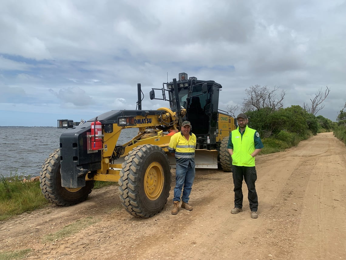 Image depicting completion of roadworks at Mitchell River Silt Jetties Reserve. Two men in high-vis jackets standing on new road with grader in background.