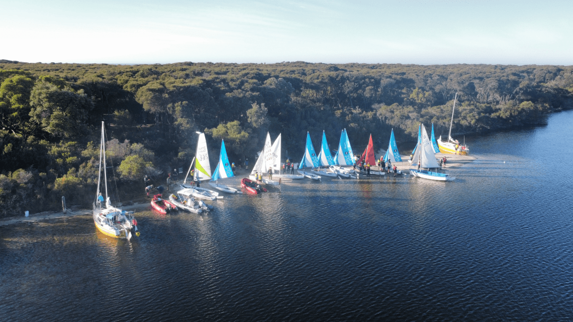 13 bright coloured sailboats lined up against the beach shoreline. A dense green bushland follows the beach