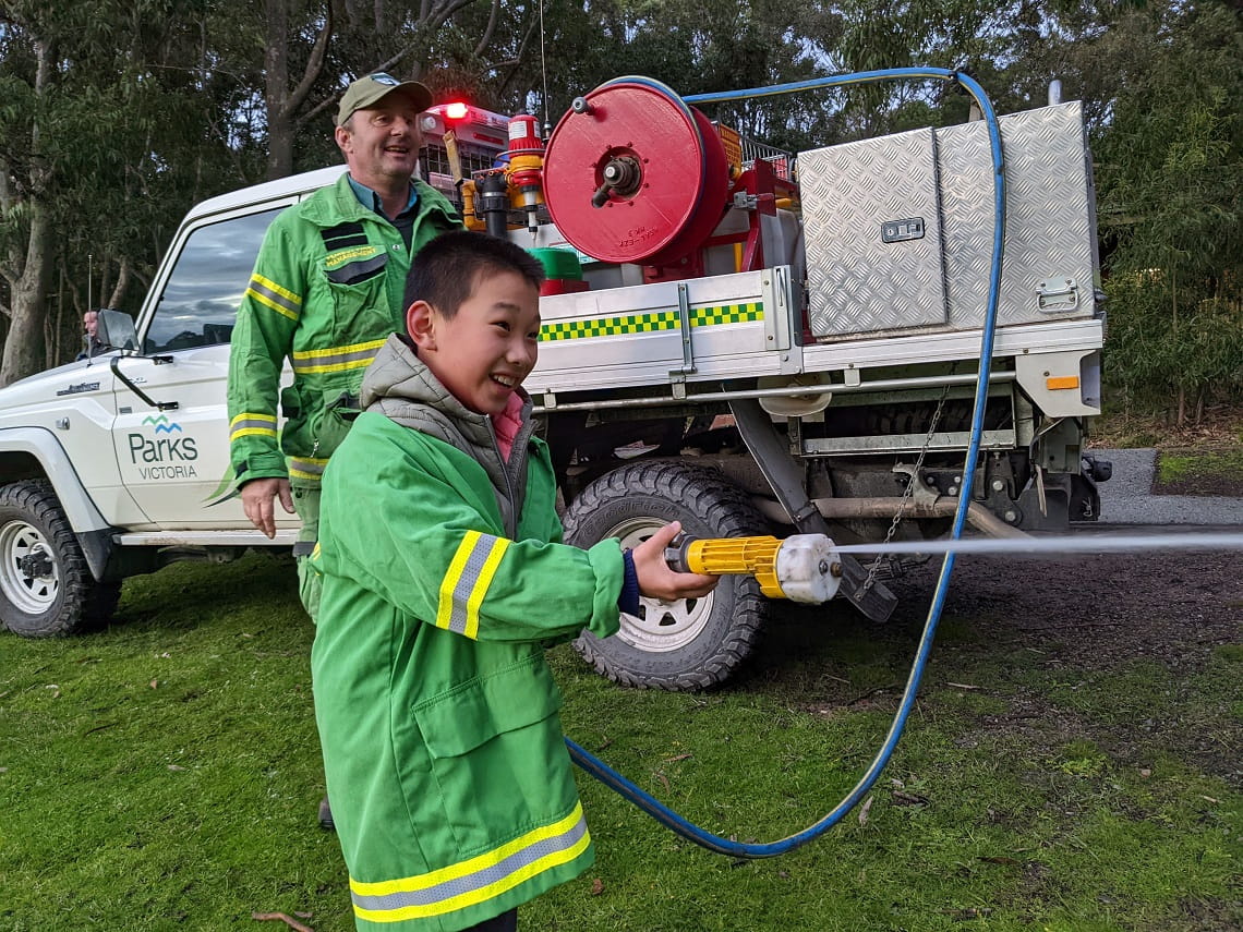 Child in PPE standing by fire truck and using the hose supervised by a ranger