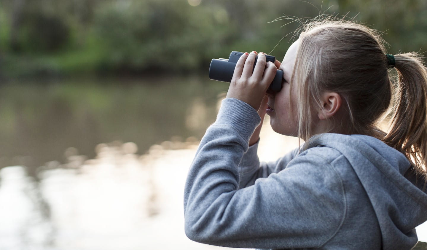 A young girl uses binoculars to search for birdlife near a river.