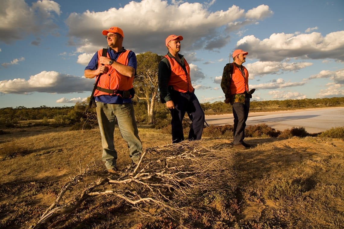 Three male Sporting Shooters volunteers stand on a dry, desert landscape, wearing orange high visibility vests and hats, with gun safely slung over their shoulders.