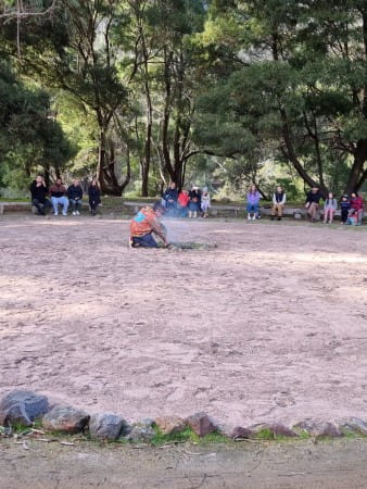 Traditional Owner crouched in centre of circle tending to smoke. Visitors are surrounding the circle watching him.