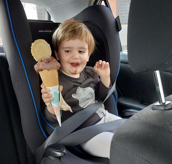 A young boy sitting in a children's car seat holding a large chocolate icecream. He is smiling with some icecream on his face