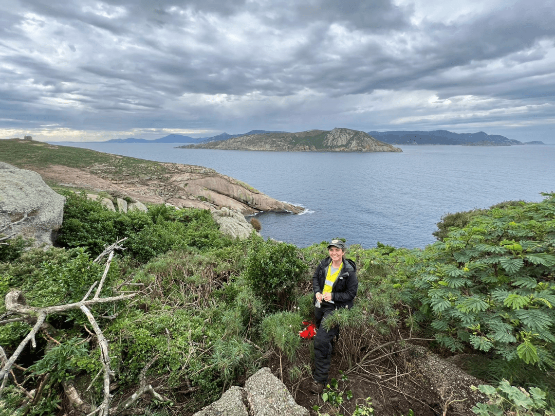 A young woman in Parks Victoria uniform stands amid dense undergrowth with sea and a rocky island in the distance behind her.