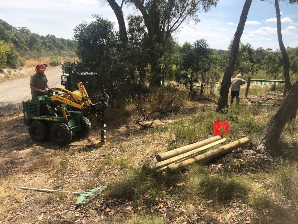 Two men in hard hats working on a four-wheeled machine with a series of logs.