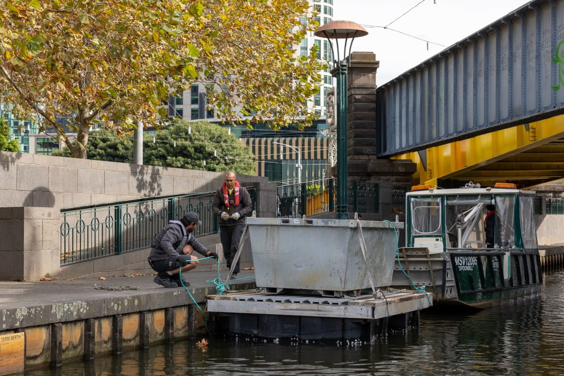 Two men in Parks Victoria uniform are unloading a skip filled with rubbish from a barge moored on the Yarra