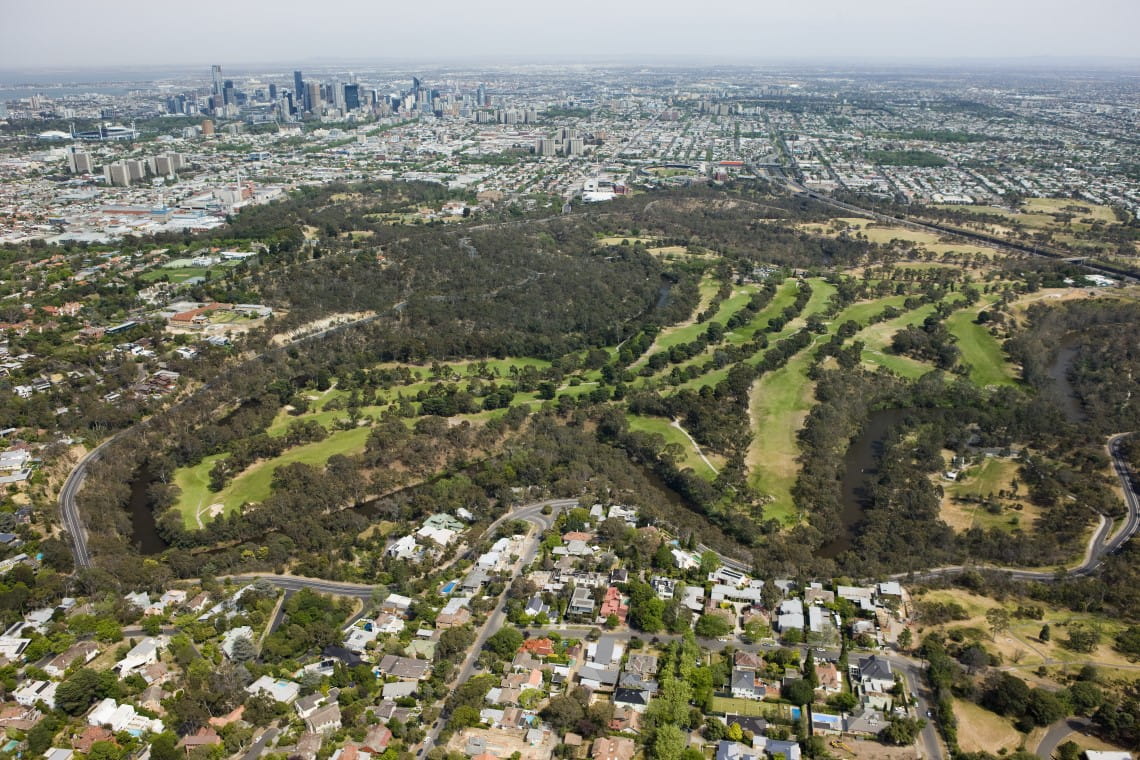 Yarra Bend Park from the air: a broad expanse of trees and grassy areas around a bend in the river, the towers of the city in the distance.