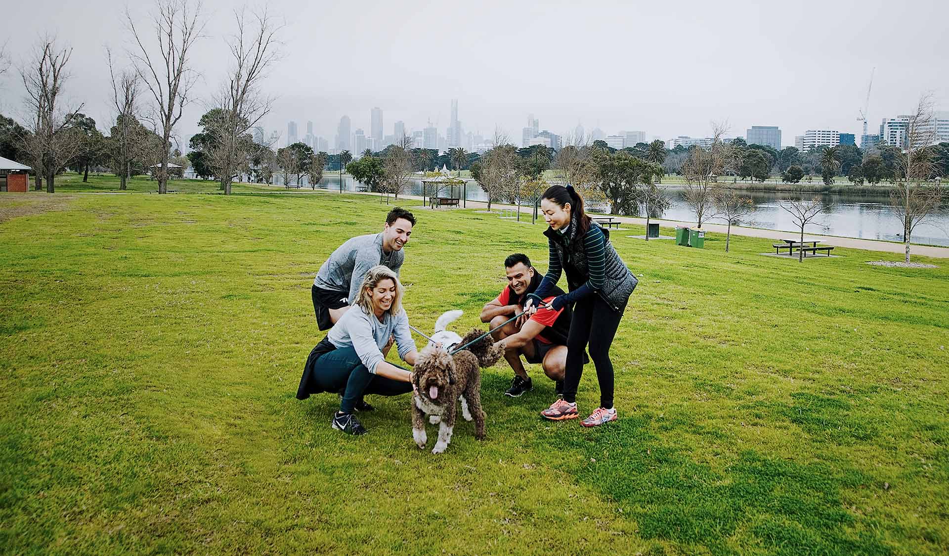Four friends pat their two dogs next to Albert Park Lake with the Melbourne CBD skyline in the background.