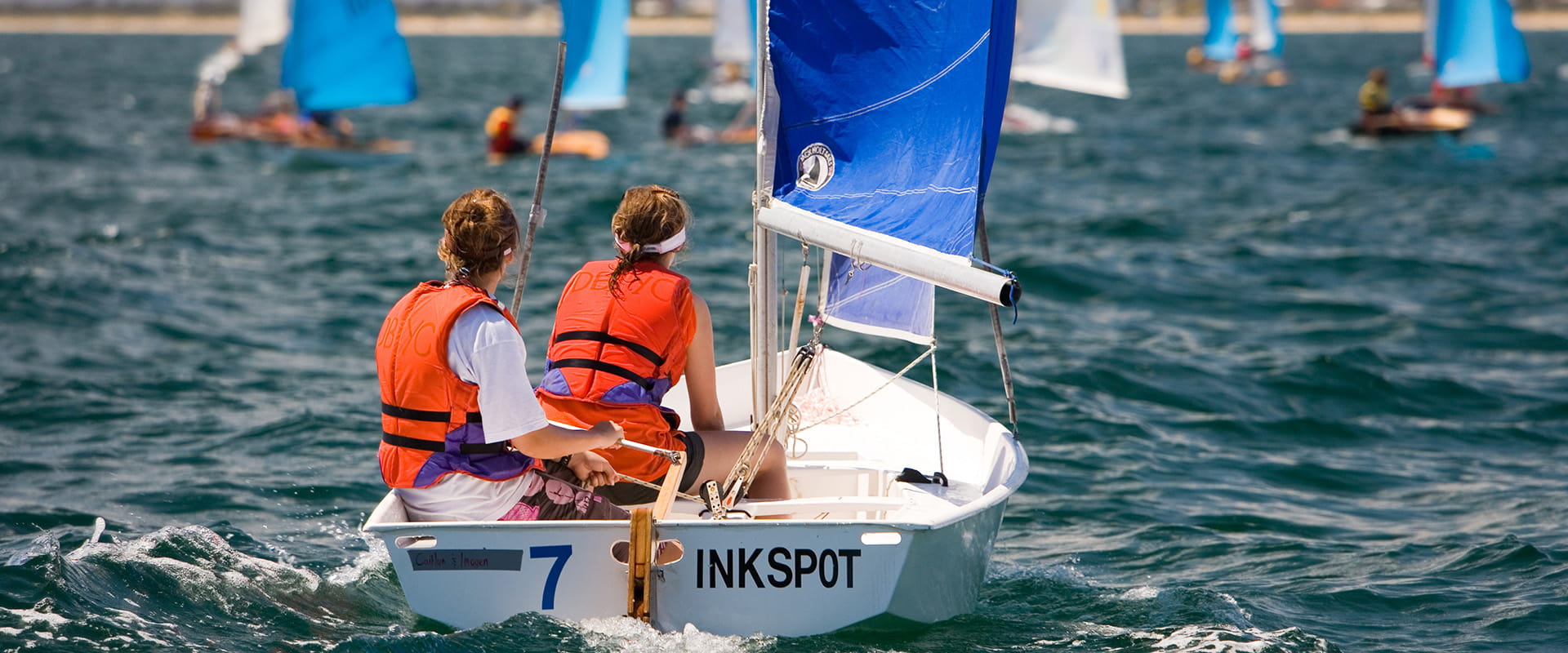 Two young women wearing bright lifejackets racing their small sailboat through the deep blue open waters.