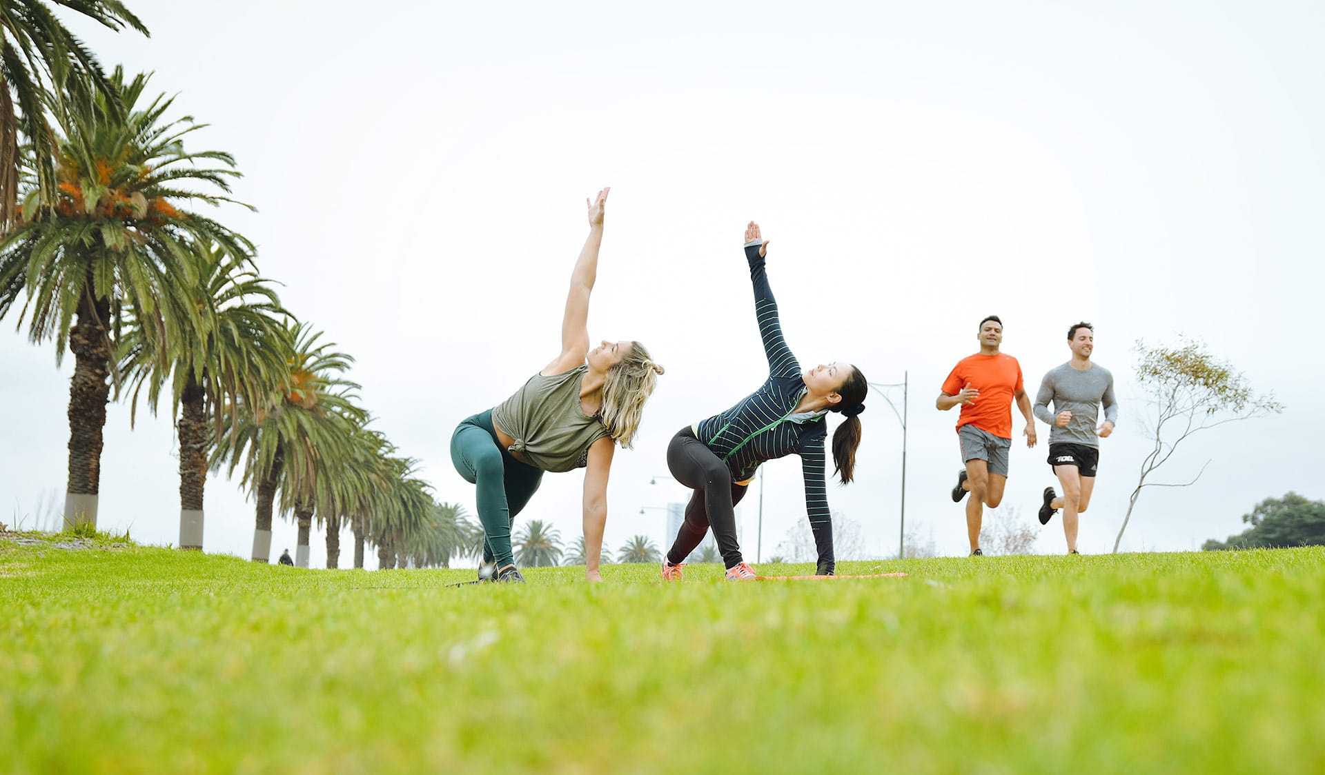 Two women practicing yoga side-by-side with two men running for fitness in the background