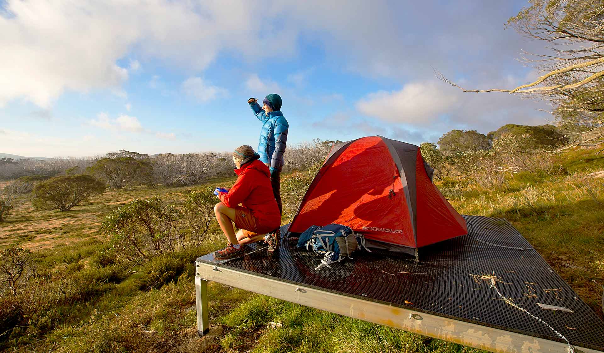Two women look out from their tent pad, upon which a red tent is setup, near Cope Hut in the Alpine National Park