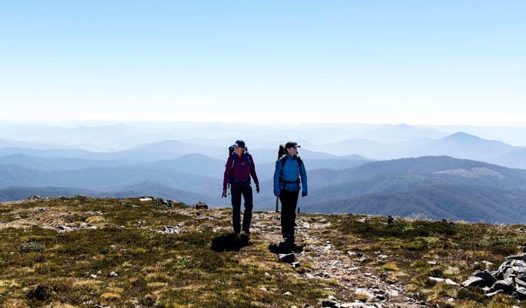 Two women hiking with a panoramic view of the mountains behind them at Alpine National Park