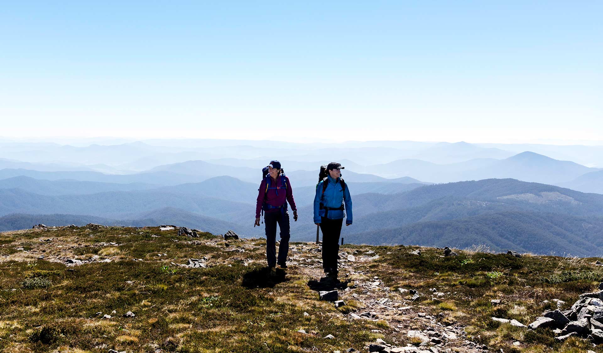 Two women hiking with a panoramic view of the mountains behind them at Alpine National Park