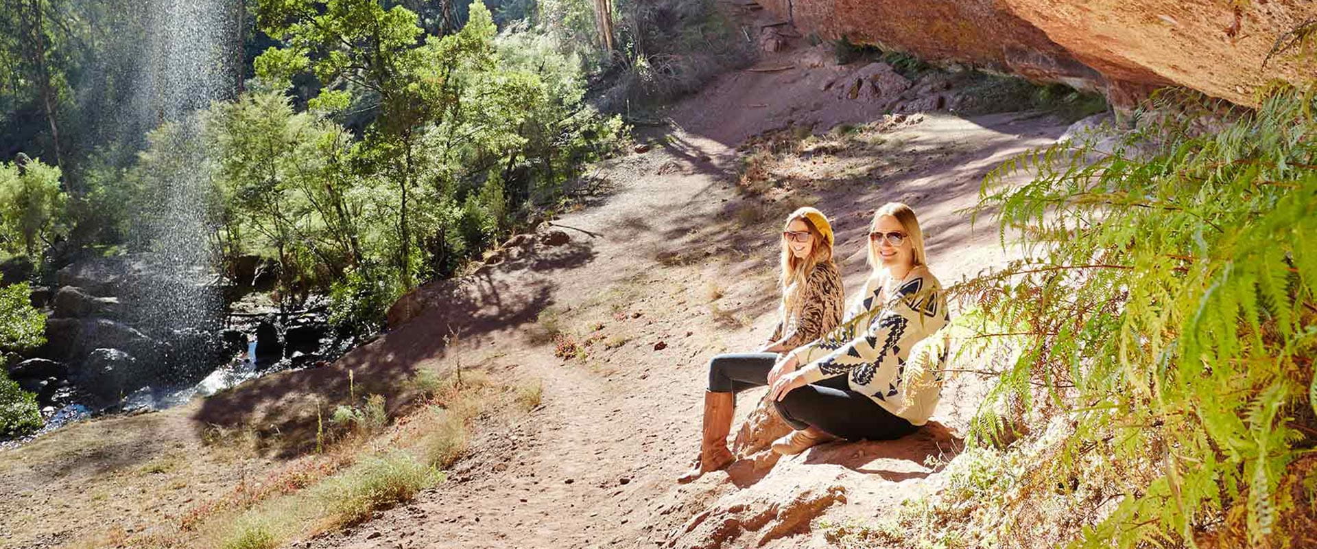Two women sit in sun next to a mountain overlooking the waterfall.