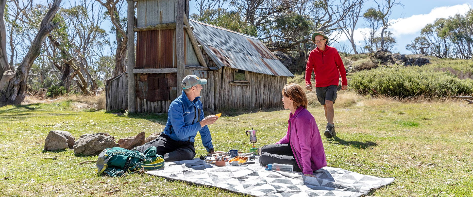 Three people enjoy a picnic out the front of a historic hut
