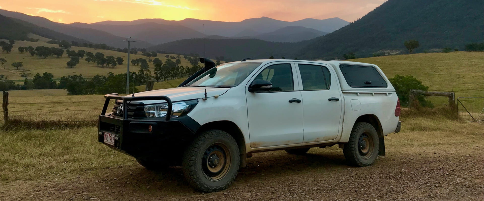 A Four Wheel Drive sits on a dirt track with a sunset highlighting the mountains in the background.