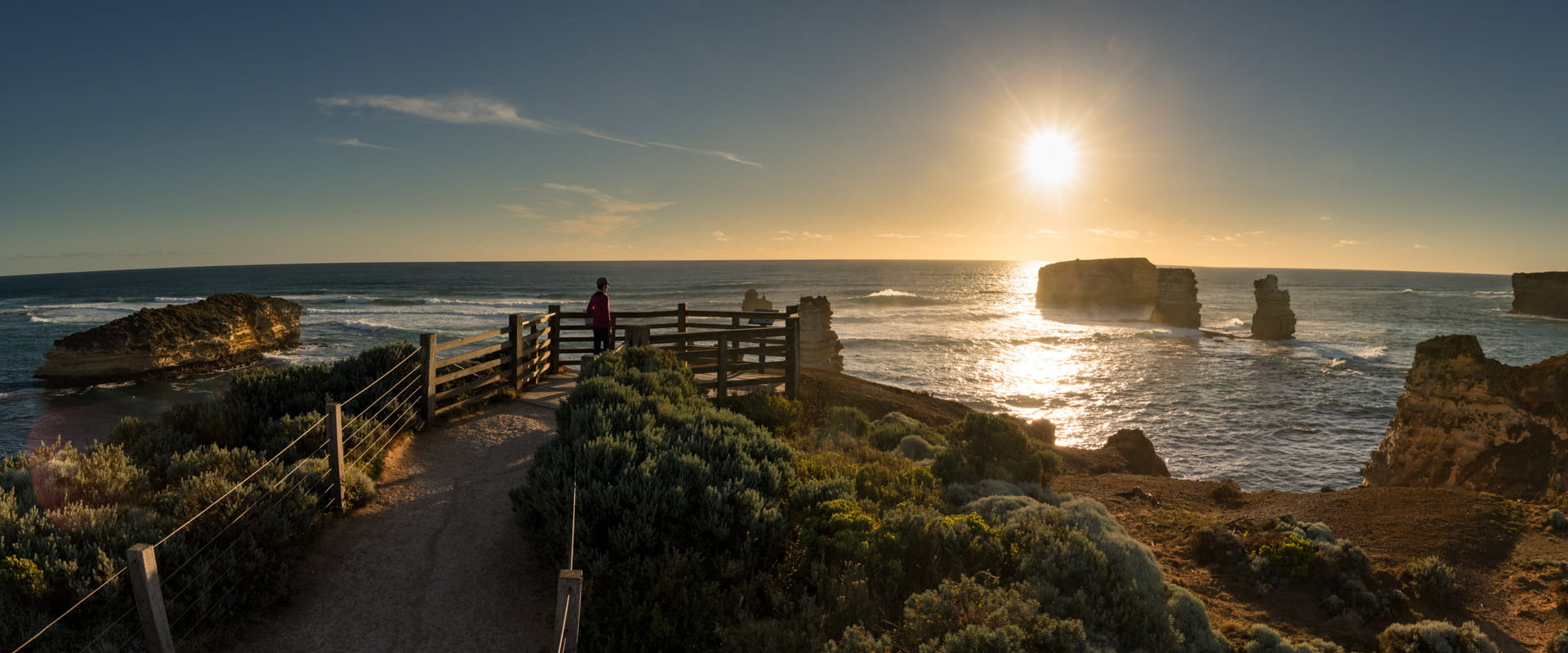 A person stands on a a viewing platform, looking out over the ocean and rock stacks. The sun sets in the background. 