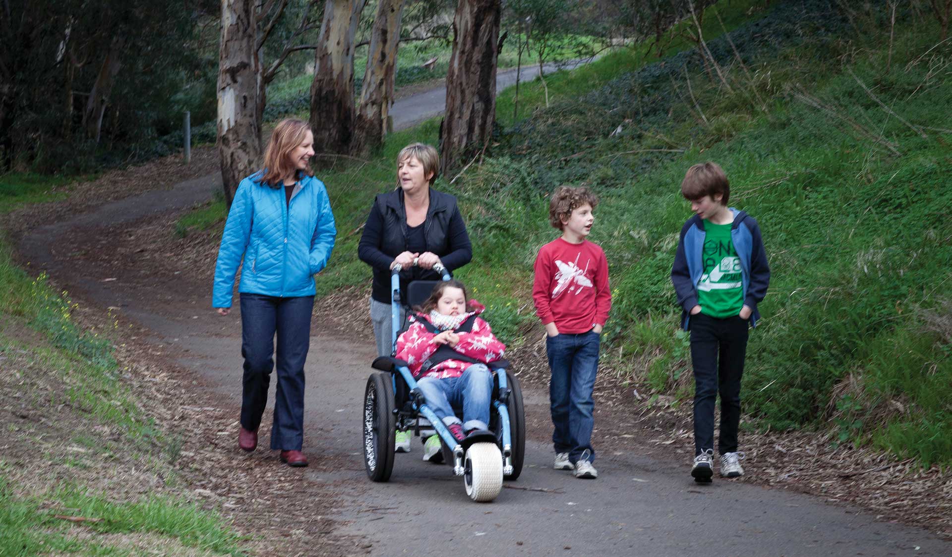 Two adults and three children, one of which is in a wheel chair, stroll along a path at Brimbank Park.