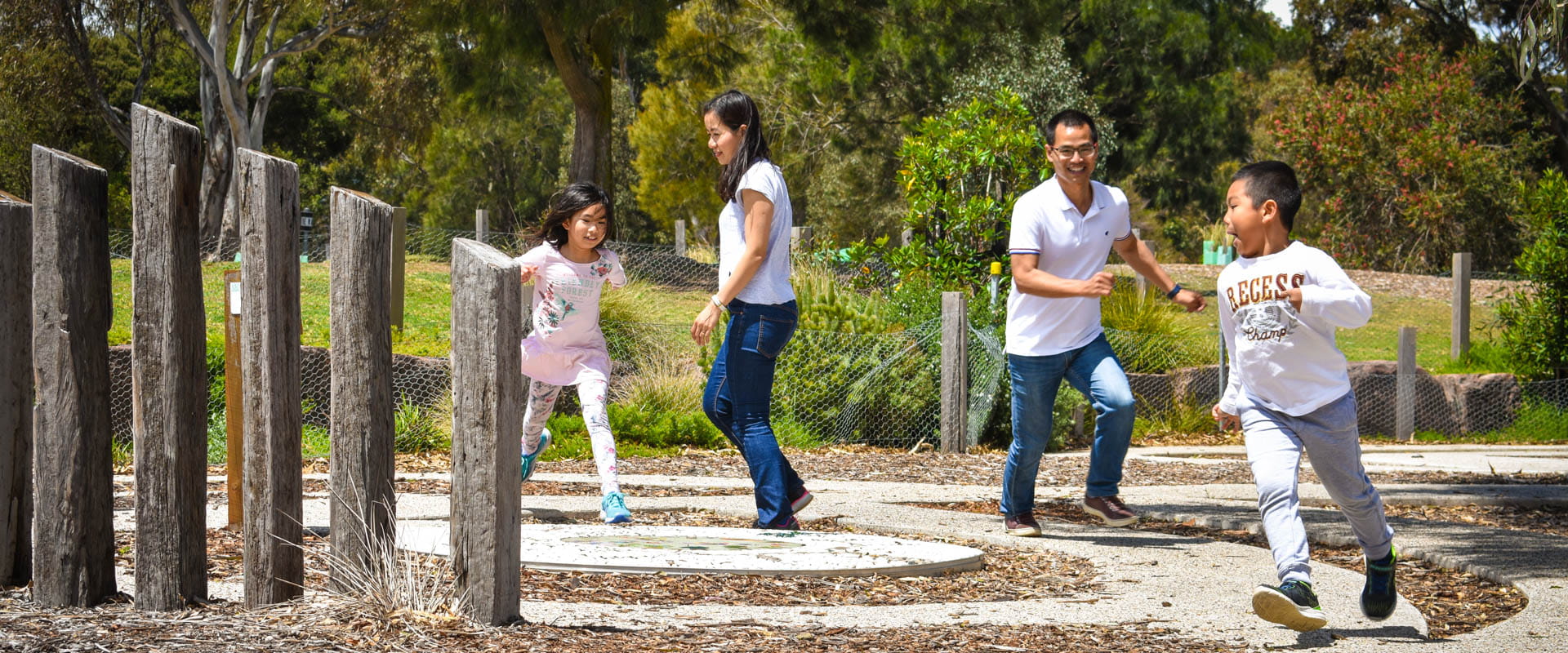 Family play and chase each other around vertical wooden logs in a paved playscape.