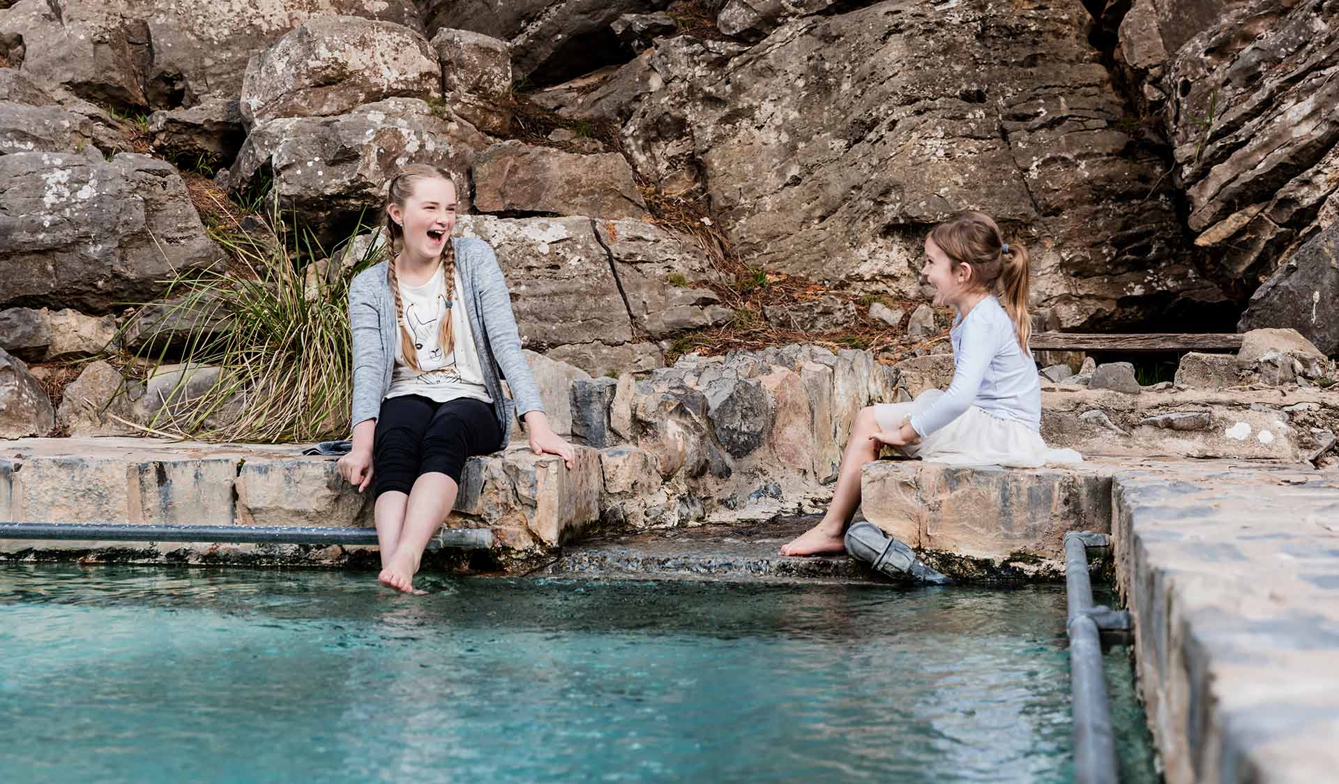 Two young girls sit on the edge of the natural spring swimming pool at Buchan Caves and dangle their feet into the water.