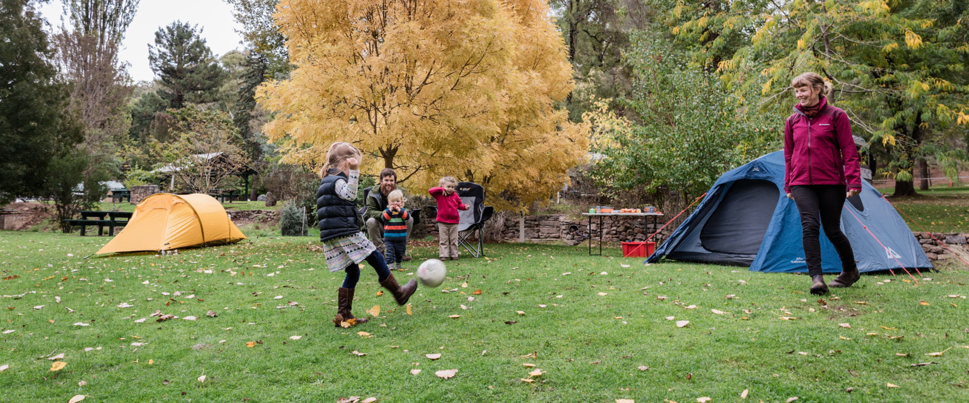 A young girl kicks a socccer ball to her mother while her family watches from their camping site amongst trees with golden and green folliage.