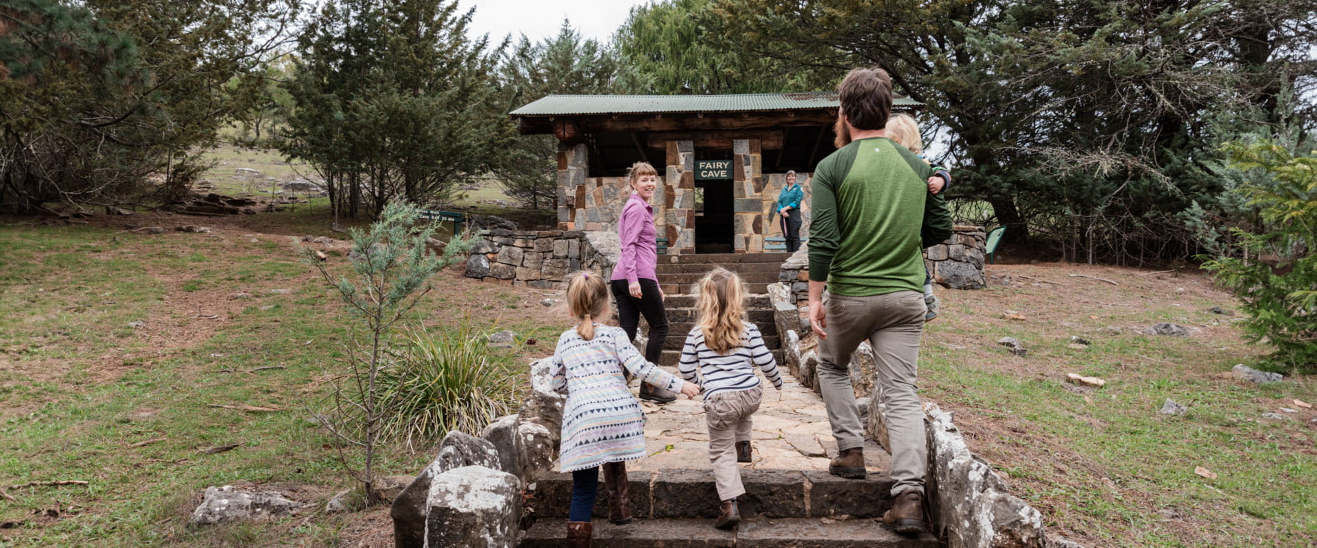 A young family walk up the rock steps and path leading to a waiting park ranger outside the stone entrance of the Fairy Cave.
