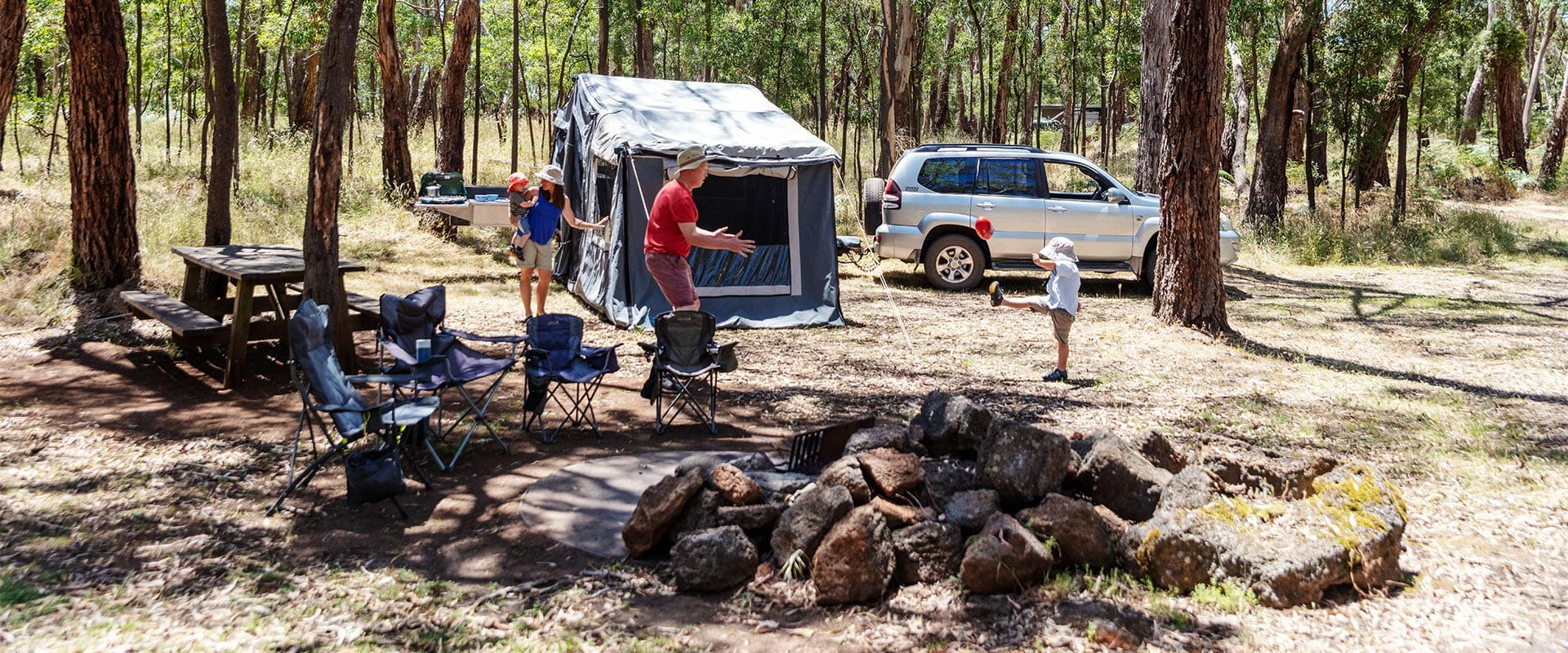 Camp chairs line an unlit fire pit in the foreground, and a man gets ready to catch a football kicked by his son while a woman holding a young child stands between a tent and picnic table with benches. A SUV towing a trailer sits behind the tent and in front of a backdrop of trees in the background.