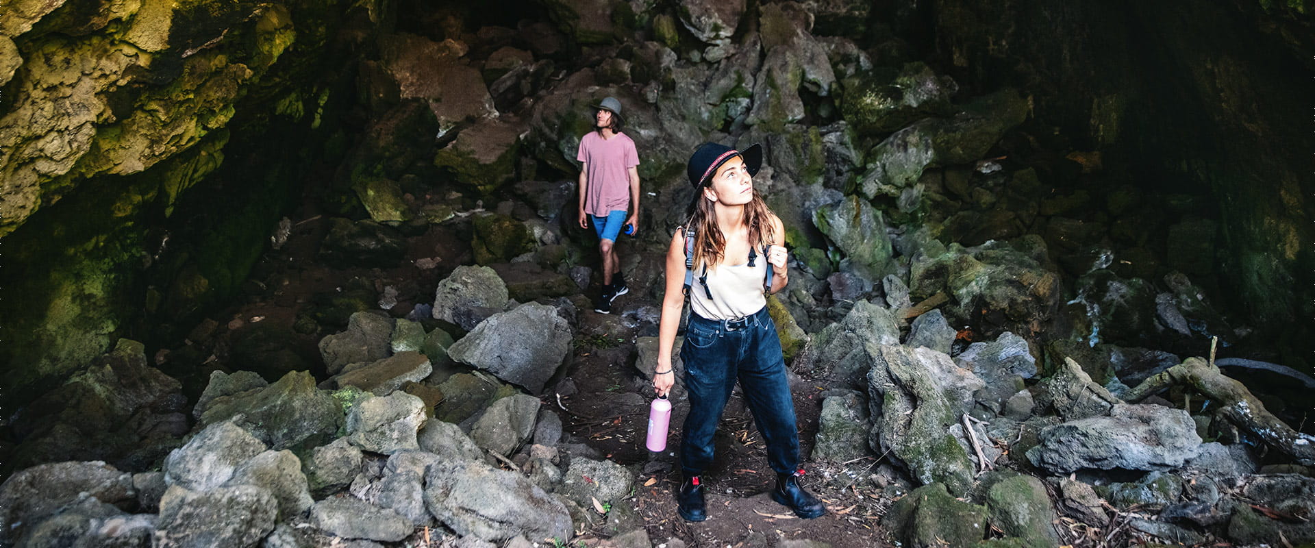 A woman stands in the foreground on a dirt path between rocks looking up, while a man looking at the cave walls follows the path in the background.