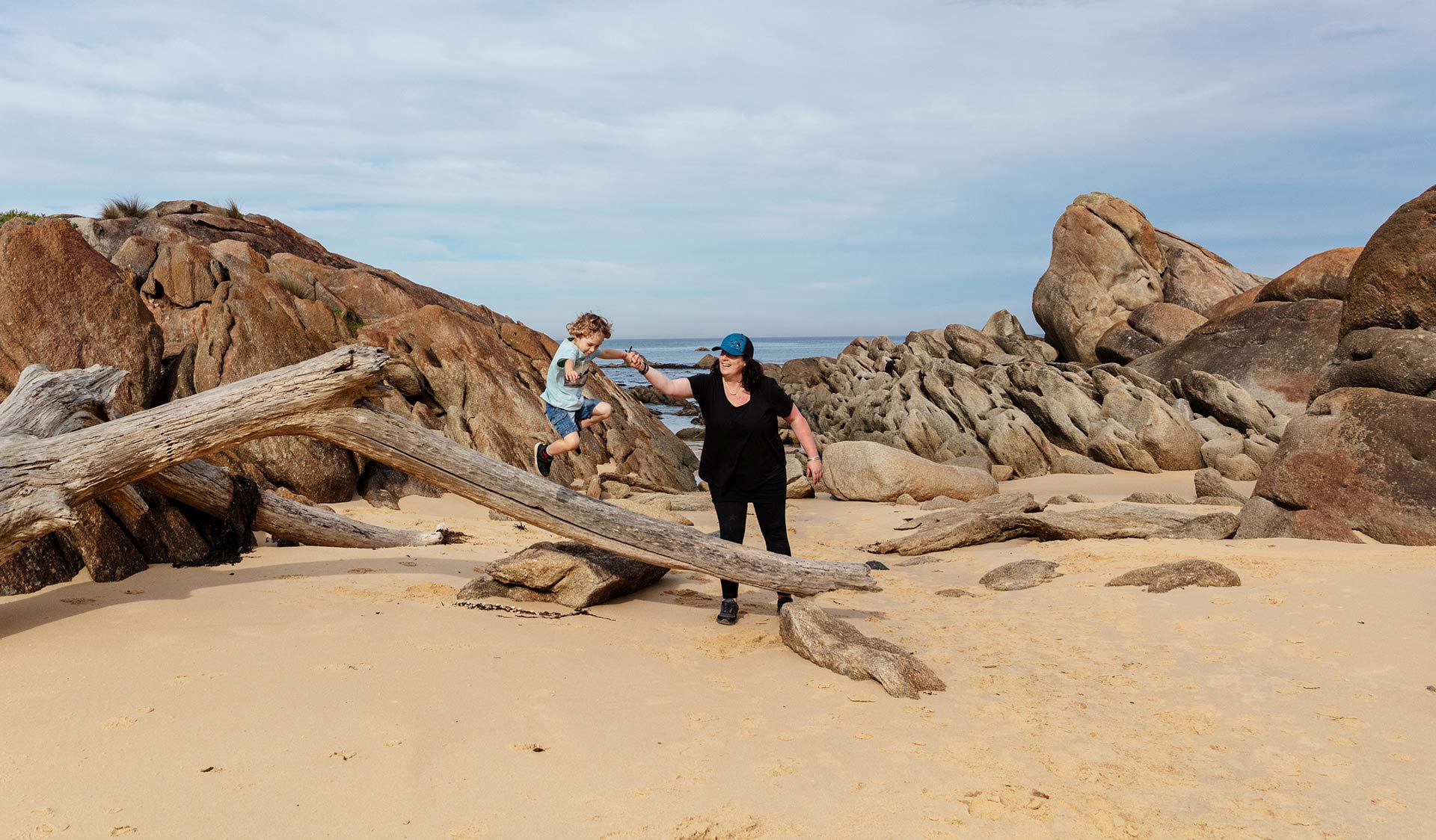 Mum helps her young son as he jumps off a large piece of drift wood at West Cape Beach.