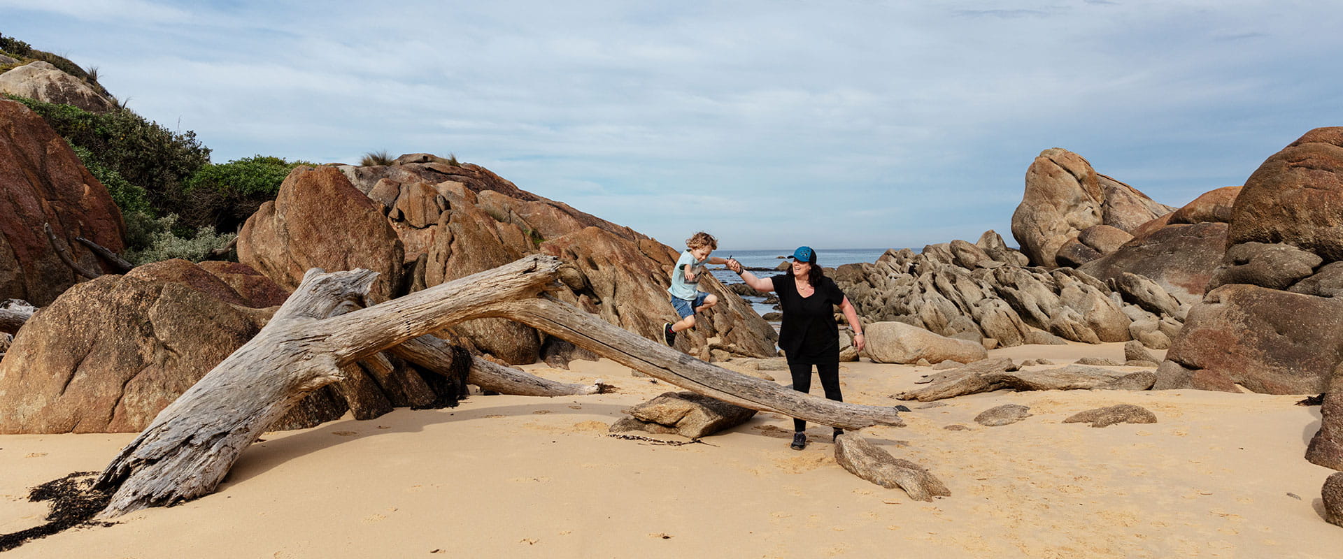 A mother wearing black clothes plays with her toddler son on a big piece of drift wood washed up on a sandy beach in front of some pink granite rocks 