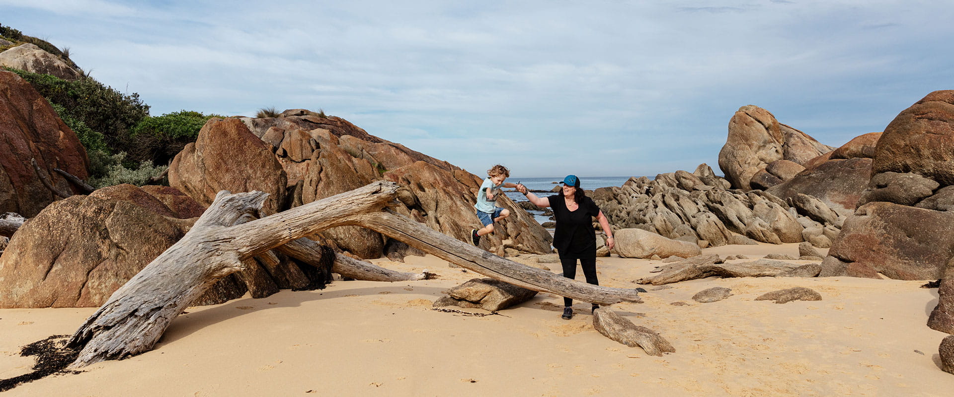 A mother wearing black clothes plays with her toddler son on a big piece of drift wood washed up on a sandy beach in front of some pink granite rocks
