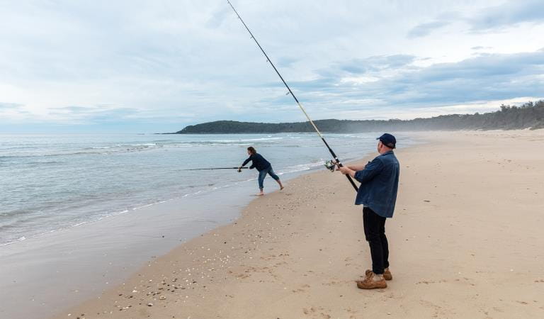 Two men cast fishing lines on a beach. 