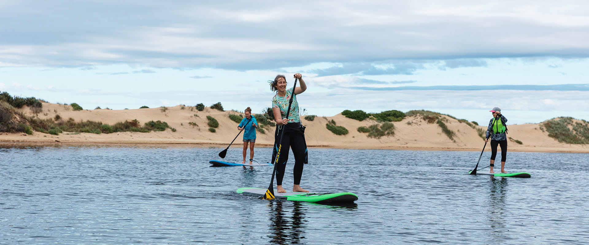 Three women stand-up-paddle board on small estuary in front of sand dunes.