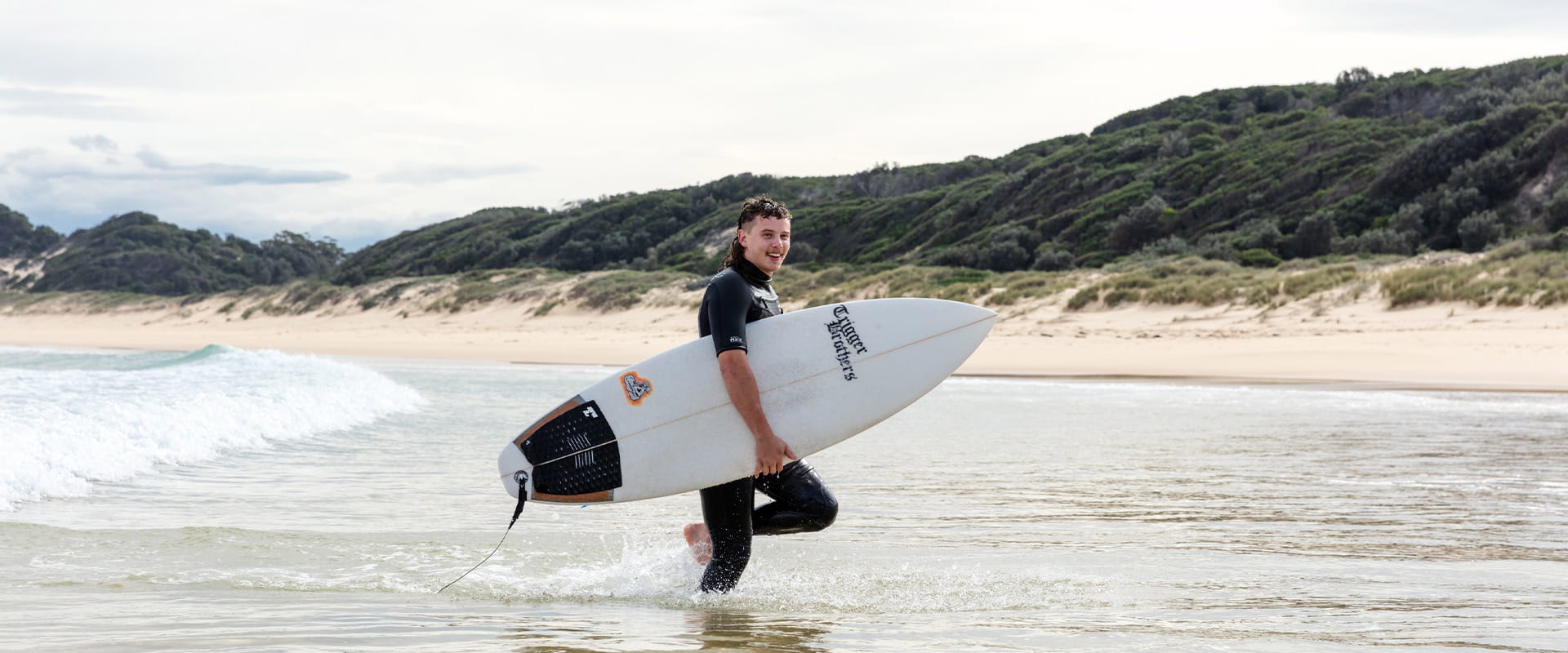 A man wearing a short sleeved wetsuit and carrying a surfboard under his right arm walks through ankle deep water towards a sandy beach with a vegetation-covered sand dunes in the background.