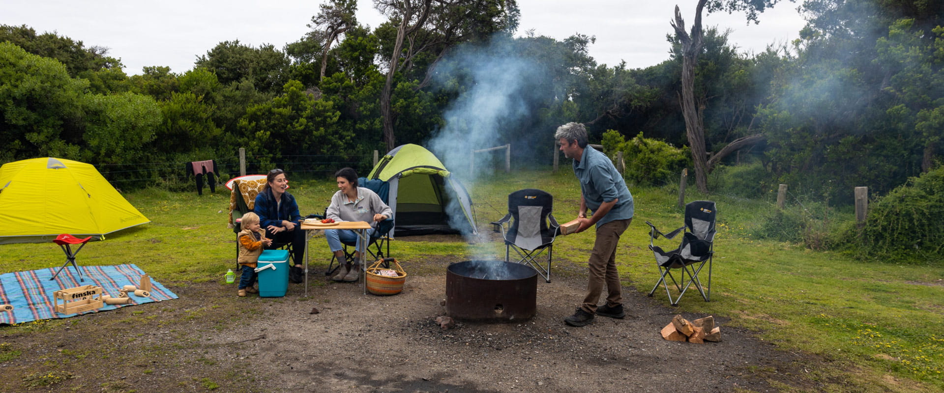 A man adds wood to a campfire while two women sit with a young child to enjoy a picnic on a small table nearby at their campsite.