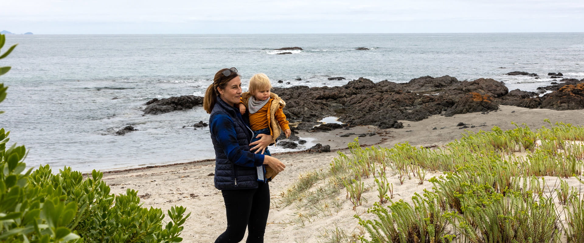 A woman holds a young child in her arms as they walk along the sandy beach with the rocky shoreline behind them.